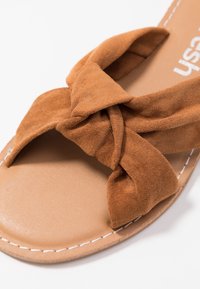 Braune Wildleder-Sandale zum Hineinschlüpfen mit einem geknoteten Design im oberen Bereich, ausgestattet mit einer tanfarbenen synthetischen Sohle und weißen Stichdetails.