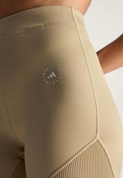 Beige tajice visoko strukirane od rastezljivog materijala s rebrastim bočnim panelom i kružnim logotipom na bedru, vidljivi detalji šivanja.