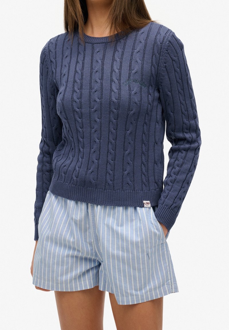 Pull en maille torsadée bleu marine avec manches longues et encolure ronde, associé à un short rayé bleu clair. Le pull présente des ourlets côtelés et un logo.