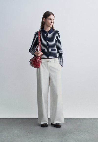Femme debout portant un cardigan à boutons rayé bleu marine et blanc, un pantalon large blanc avec ceinture, des chaussures noires, et tenant un sac à main rouge sur l'épaule.