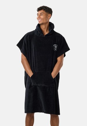 Homme portant un poncho serviette noir oversize avec capuche, poche avant et logo ancre blanc, souriant et regardant vers la droite.