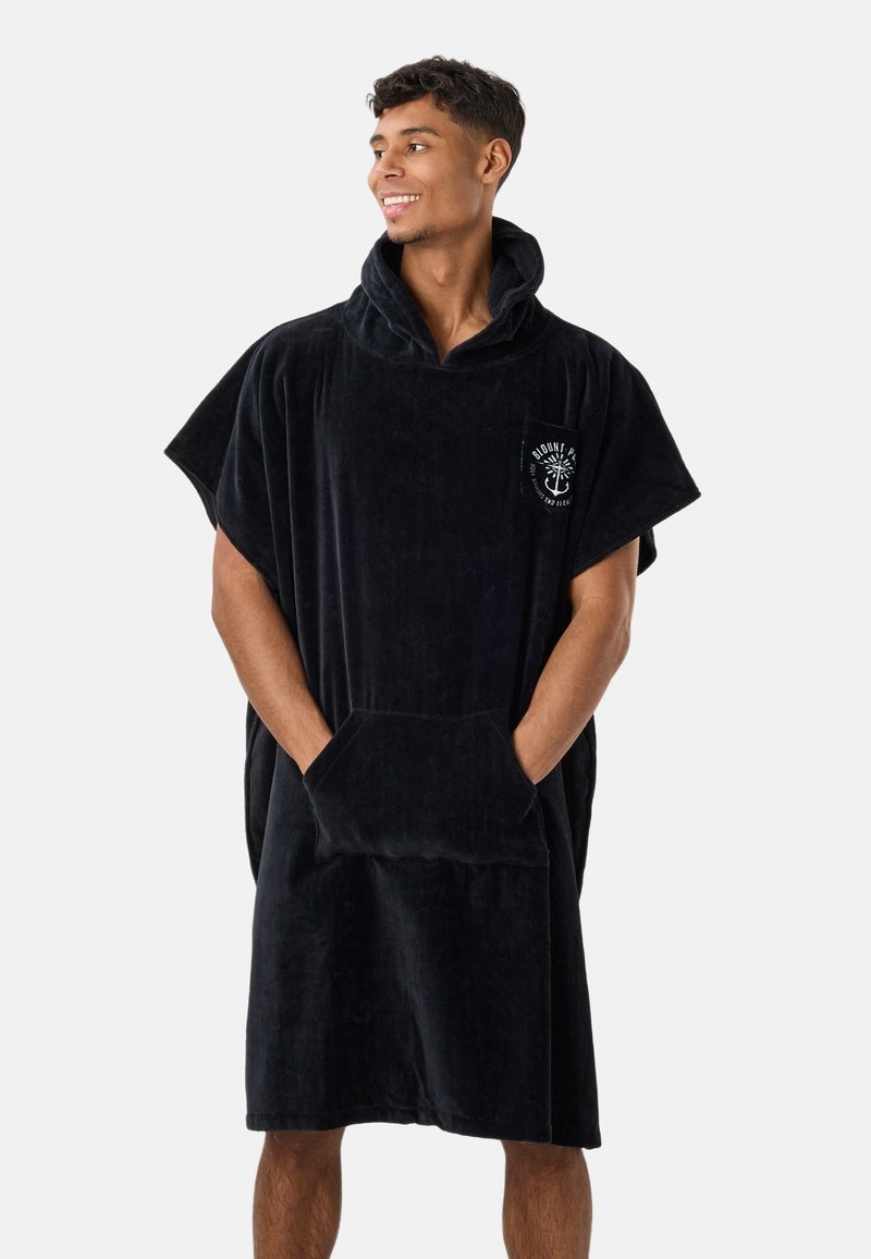 Άνδρας που φοράει μαύρο μεγάλο πανωφόρι-poncho με κουκούλα και μπροστινή τσέπη, με λευκό λογότυπο άγκυρας, χαμογελά και κοιτάζει δεξιά.