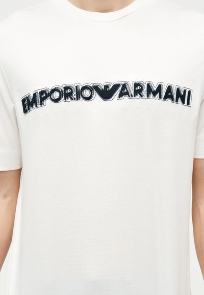 Λευκό μπλουζάκι με μαύρο κεντητό λογότυπο "EMPORIO ARMANI" στο στήθος και μικρό τμήμα των χεριών του φορέα ορατό.