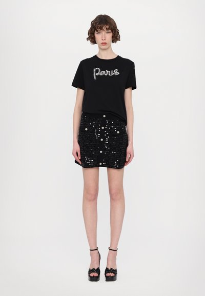 Schwarzes T-Shirt mit Kristallschriftzug "Paris", kombiniert mit einem schwarzen Rock aus Pailletten mit Taschen und dekorativen Knöpfen. Schwarze Absatzsandalen.