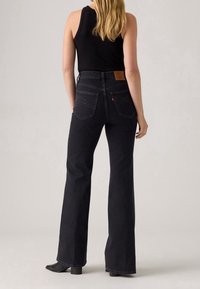 Femme aux longs cheveux blonds portant un haut noir sans manches et un jean noir taille haute avec jambes évasées, debout devant un fond uni.