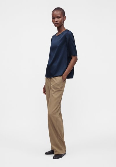 Femme debout portant un chemisier bleu foncé à manches courtes, un pantalon beige large et des chaussures noires sur un fond clair uni.