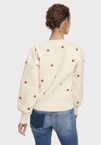 Sweatshirt beige clair avec broderie de araignée rouge, doté d'un col rond, de manches longues bouffantes et d'une finition côtelée. Assorti avec un jean bleu.