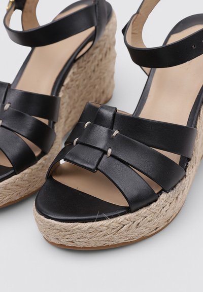 Sandalias de cuña de cuero negro con suelas de plataforma de yute trenzado y correas en el tobillo sobre un fondo blanco.