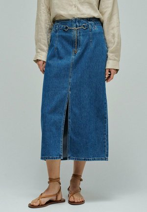 Falda midi de denim con abertura frontal, detalle de cinturón de cadena, combinada con camisa beige de manga larga y sandalias planas marrones con tiras.