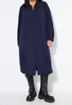 Femme portant une robe bleu marine à manches longues boutonnée avec des bottes noires épaisses montant jusqu'aux genoux, debout sur un fond blanc.