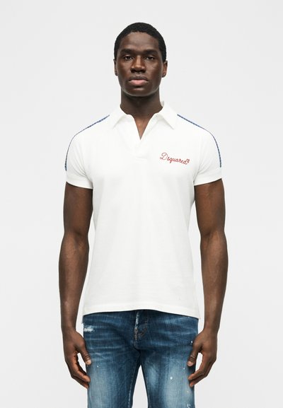 Homme portant un polo blanc à manches courtes avec le logo rouge "Dsquared²" et des détails à rayures bleues sur les épaules, associé à un jean bleu déchiré.