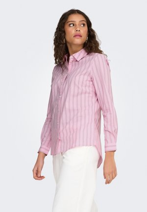 Kortsude juustega naine, kandes roosade triipudega nööbitavat särki ja valgeid pükse, vaadates läände tavalise tausta ees.