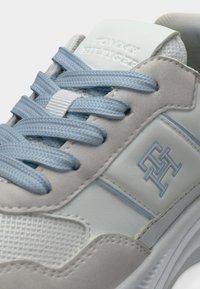 Ανοιχτόγκρι αθλητικό παπούτσι με υφή από πλεκτό δίχτυ και λείες επιφάνειες, με τόνους από γαλάζια κορδόνια, που διαθέτει ένα διακριτικό ανάγλυφο λογότυπο στη γλώσσα.