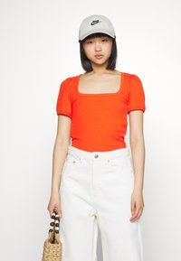 Camisola curta de manga com pregas, em laranja brilhante e decote quadrado, combinada com calças largas brancas e uma bolsa de palha. Boné cinza da Nike usado.