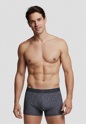 Man zonder shirt met kort bruin haar, die donkere grijze boxershort draagt met kleine lichte verenpatronen, staand tegen een effen grijze achtergrond.