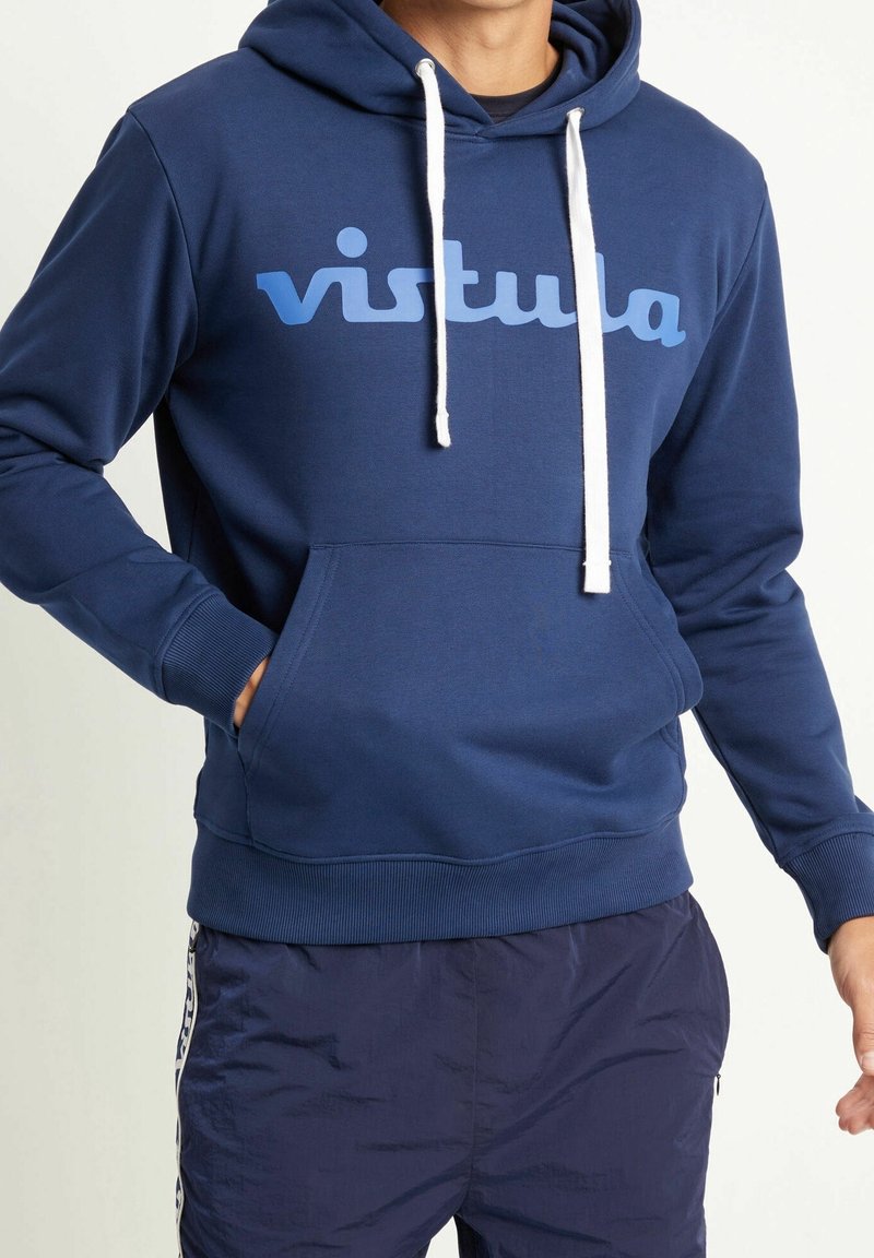 Homme portant un sweat à capuche bleu marine avec cordons blancs et logo "vistula", associé à un pantalon de survêtement bleu foncé avec une bande blanche sur le côté.