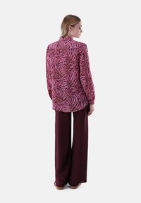 Blusa a maniche lunghe in rosa e bordeaux con un motivo a spirale, abbinata a pantaloni bordeaux a gamba larga. Tessuto morbido e vestibilità rilassata.