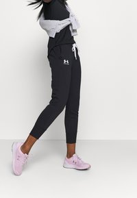 Schwarze Sporthosen mit weißen Kordelzügen und Logo, kombiniert mit einem schwarzen T-Shirt und einem grauen Pullover, der um die Taille gebunden ist. Dazu werden rosa Turnschuhe getragen.