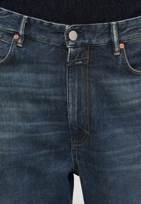 Jeans din denim de culoare albastru închis, cu croială dreaptă, având nituri din cupru, cusături portocalii și o etichetă pe brâu.