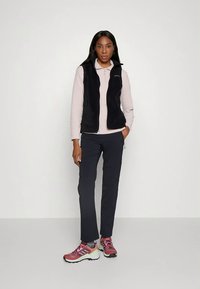 Μαύρο γιλέκο από φleece πάνω από ρόδινο μακρυμάνικο μπλουζάκι, συνδυασμένο με σκούρα παντελόνια και πολύχρωπα αθλητικά παπούτσια. Απλός σχεδιασμός και άνετη εφαρμογή.