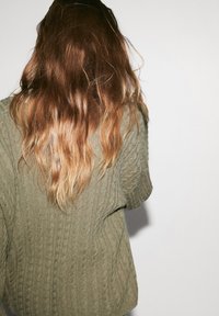 Olive green knitted sweater with a textured cable pattern, featuring a loose fit and wide sleeves, worn by a person with wavy hair.