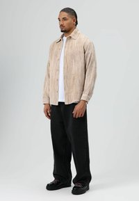 Lichtbeige, geribde overhemd met knopen en kraag, gedragen over een wit T-shirt. Gecombineerd met losse zwarte broek en zwarte veterschoenen.