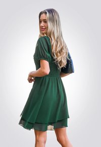 Vestido verde, de longitud hasta la rodilla, con mangas abullonadas y cintura fruncida. Falda de capas transparentes en una tela ligera, combinada con un pequeño bolso negro.