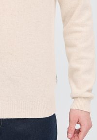 Beige, lange mouwen, gebreide trui met geribde manchetten, gedragen met donkere denim jeans, focus op de onderkant van de torso en rechterarm.
