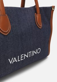 Mörkblå denim totebag med bruna läderhandtag och detaljer, som har vit "VALENTINO" logotext på framsidan.