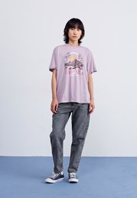 Lilac graphic t-shirt with mountain design and "ROXY" text, paired with gray jeans and navy sneakers. Short sleeves, relaxed fit.