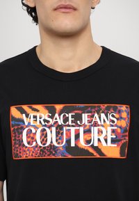 Black cotton t-shirt featuring a colorful graphic print with orange and blue accents, displaying the text "VERSACE JEANS COUTURE."