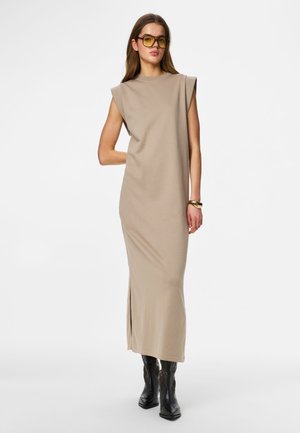 Femeie purtând o rochie maxi bej fără mâneci, cizme de cowboy negre, ochelari de soare cu lentile galbene și o brățară aurie, stând în fața unui fundal simplu.