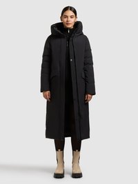 Abrigo largo negro tipo puffer con capucha, con bolsillos laterales, cierre de cremallera y superficie texturizada, combinado con botines beige hasta el tobillo.