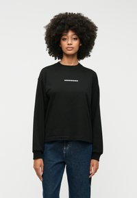 Melna garā piedurkņu džemperis ar baltiem uzrakstiem "MINDWORK". Iezīmē apaļu kaklu, brīvu piegriezumu un mīkstu kokvilnas materiālu. Apvienots ar ziliem džinsiem.