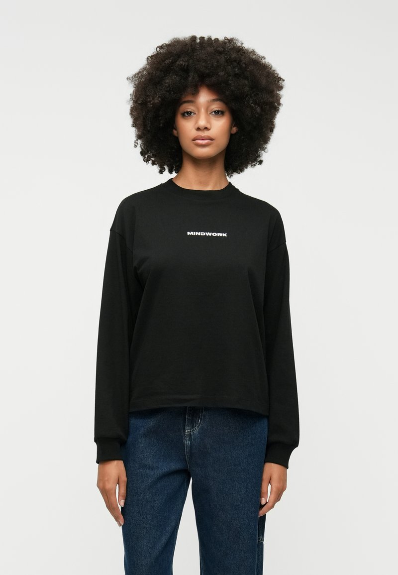 Schwarzer Sweatshirt mit langen Ärmeln und "MINDWORK" in Weiß bedruckt. Mit Rundhalsausschnitt, lockerer Passform und weichem Baumwollmaterial. Kombiniert mit blauen Jeans.