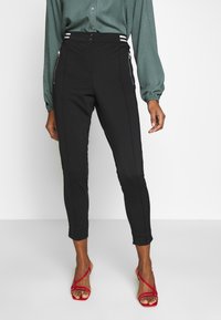 Svarta skräddarsydda byxor med hög midja och silverfärgade dragkedjefickor, tillsammans med en grön texturerad blus och röda sandalettklackar.