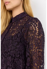 Kvinna som bär en mörklila blus i spets med blommönster och krage, där den genomskinliga spetsen syns på axel och arm.