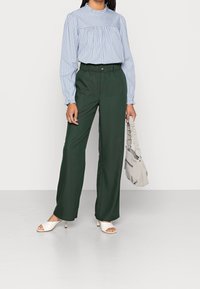 Pantalon large vert avec taille élastique, associé à un chemisier à manches longues bleu clair rayé. Sandales blanches à talons ouvertes. Sac gris.