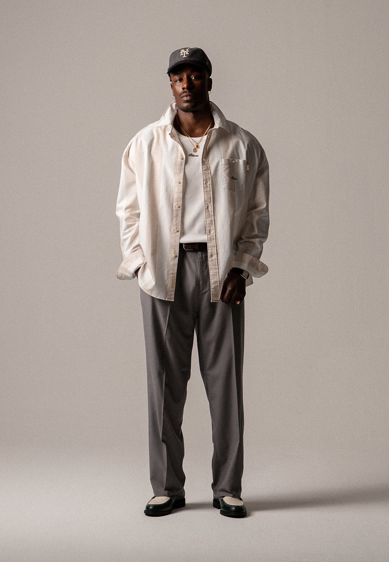 Light beige oversized shirt, white t-shirt, grey trousers, black and white shoes, dark baseball cap, gold necklace, minimalistic accessories.