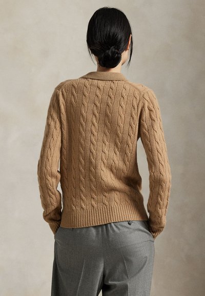 Fekete hajú nő kontyban, barna kötött mintás pulóvert és szürke nadrágot visel, zsebében tartja a kezét, egy egyszínű háttér előtt áll.