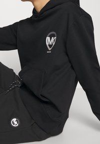 Black hooded sweatshirt, cotton blend, features a front pocket, gray logo on chest and matching logo on pants with patterned drawstrings.