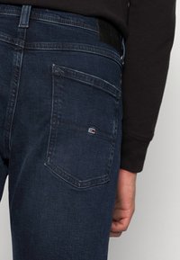 Vaqueros de mezclilla azul oscuro con un pequeño parche de logo en el bolsillo trasero. Presentan costuras estándar y un acabado texturizado.