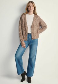 Sweter kardigan w jasnobrązowym kolorze z teksturowanym wzorem, charakteryzujący się luźnym krojem, otwartym przodem i wyraźnymi guzikami, zestawiony z niebieskimi spodniami o szerokich nogawkach.