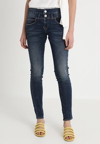 Smala mörka denimjeans med medelhög midja, två framfickor och två knappstängningar. Bärs med gula sandaler med remmar.