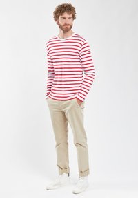 Camicia a maniche lunghe rossa e bianca a righe, in cotone; abbinata a pantaloni beige e sneakers bianche. Design a taglio dritto e casual.