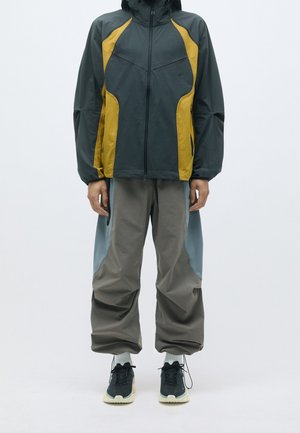 Persoon draagt een zwart- en mosterdkleurig jack met rits, grijze broek met lichtblauwe panelen, witte sokken en zwarte sneakers met witte zolen.
