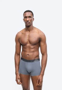 Lihaksikas mies seisoo harmaat Calvin Klein -bokserialushousut päällään ja katsoo suoraan eteenpäin vaaleanharmaata taustaa vasten.