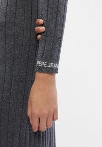 Siva rebrasta pletenina na rokavu s belim logotipom "PEPE JEANS" na manšeti. Roka drži rukav, prikazujoc teksturo in dizajn.