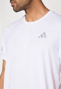 Bílé sportovní tričko s krátkým rukávem a hladkou texturou, s reflexním šedým logem na hrudi a horních rukávech.