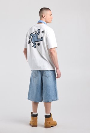 Jeune homme portant un t-shirt blanc avec un graphique d'une silhouette dansante bleue, un short en denim bleu clair, des bottes beige et une écharpe bleue, debout de profil.